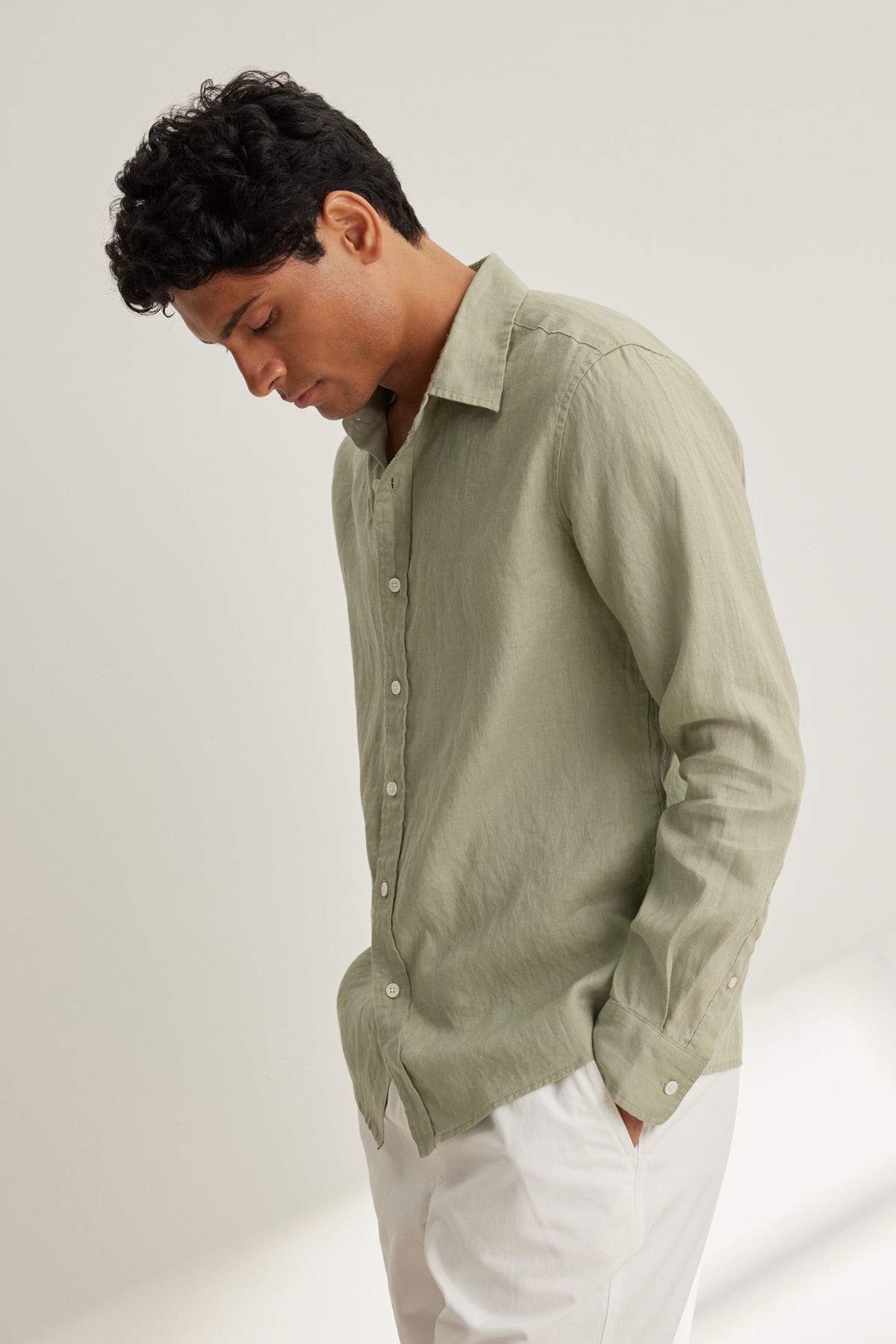  A man stands with hands in pockets, looking down against a neutral backdrop, wearing the CARUSO LINEN BUTTON-UP SHIRT by Velvet by Graham & Spencer in light green paired with white pants. 