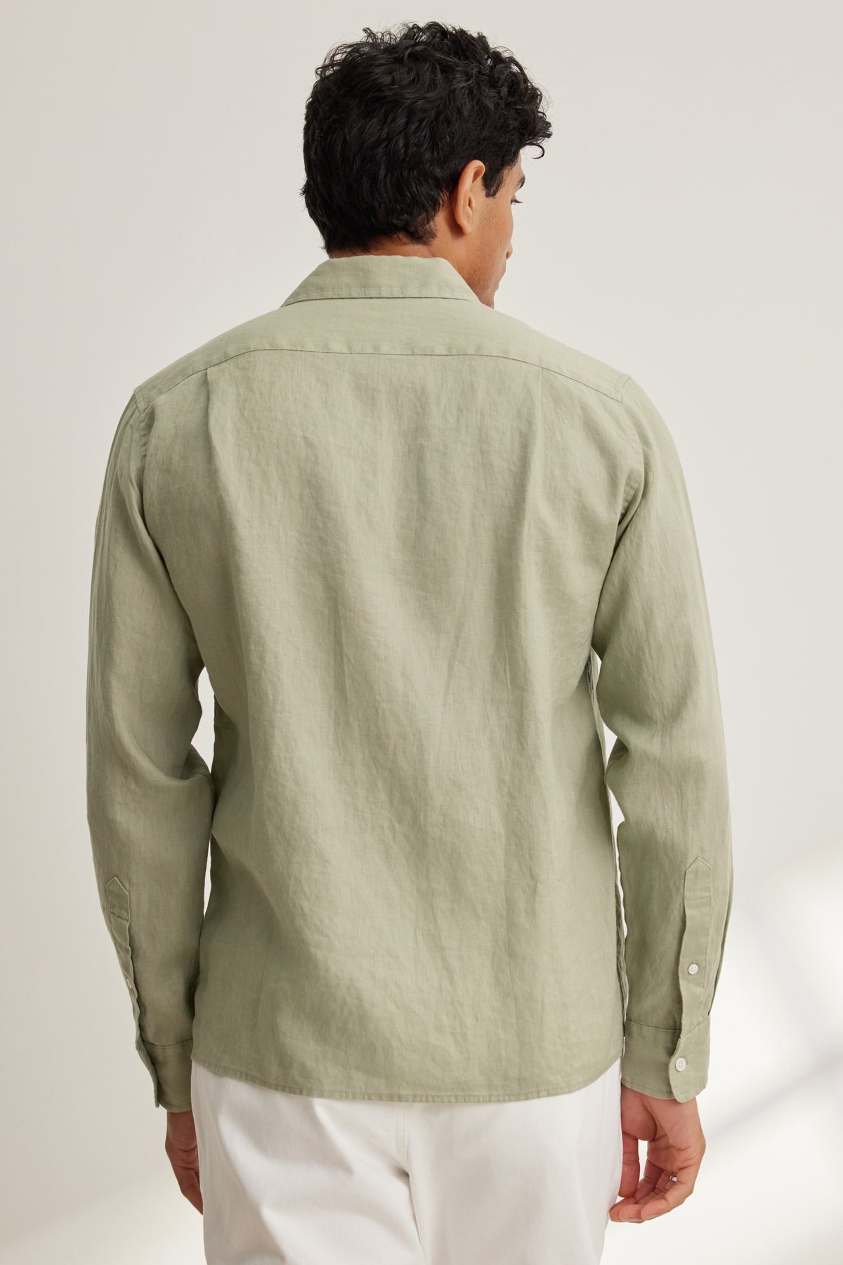   A man stands with his back to the camera, wearing the Velvet by Graham & Spencer CARUSO LINEN BUTTON-UP SHIRT in light green and white pants. 
