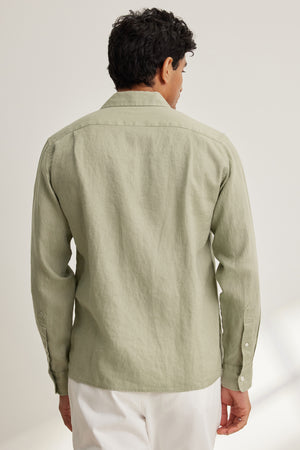 A man stands with his back to the camera, wearing the Velvet by Graham & Spencer CARUSO LINEN BUTTON-UP SHIRT in light green and white pants.