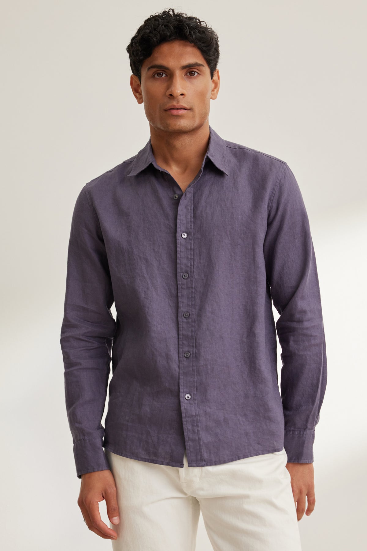   A man faces forward against a plain light background, wearing the Velvet by Graham & Spencer CARUSO LINEN BUTTON-UP SHIRT in purple with long sleeves and white pants. 
