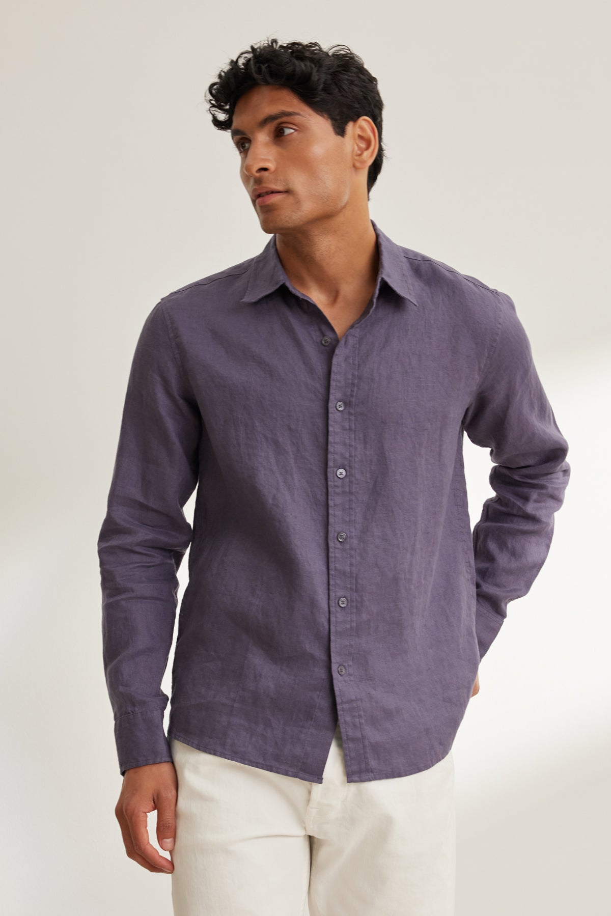 A man wears the CARUSO LINEN BUTTON-UP SHIRT by Velvet by Graham & Spencer in purple, paired with white pants, standing against a plain light background.-39152342794433