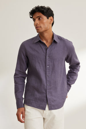 A man wears the CARUSO LINEN BUTTON-UP SHIRT by Velvet by Graham & Spencer in purple, paired with white pants, standing against a plain light background.