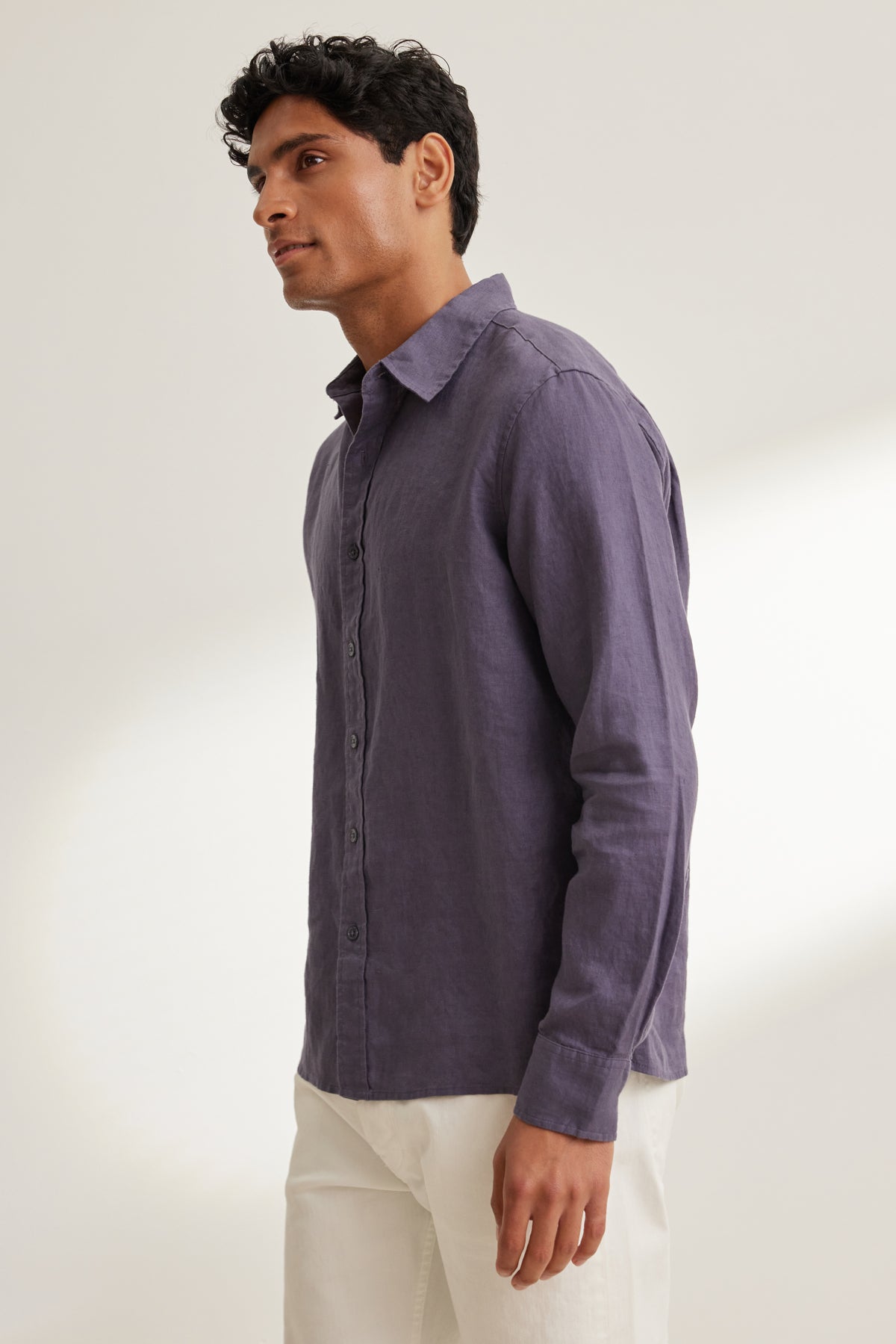 A man stands sideways against a plain background, wearing the Velvet by Graham & Spencer CARUSO LINEN BUTTON-UP SHIRT in purple with white pants.-39152342761665