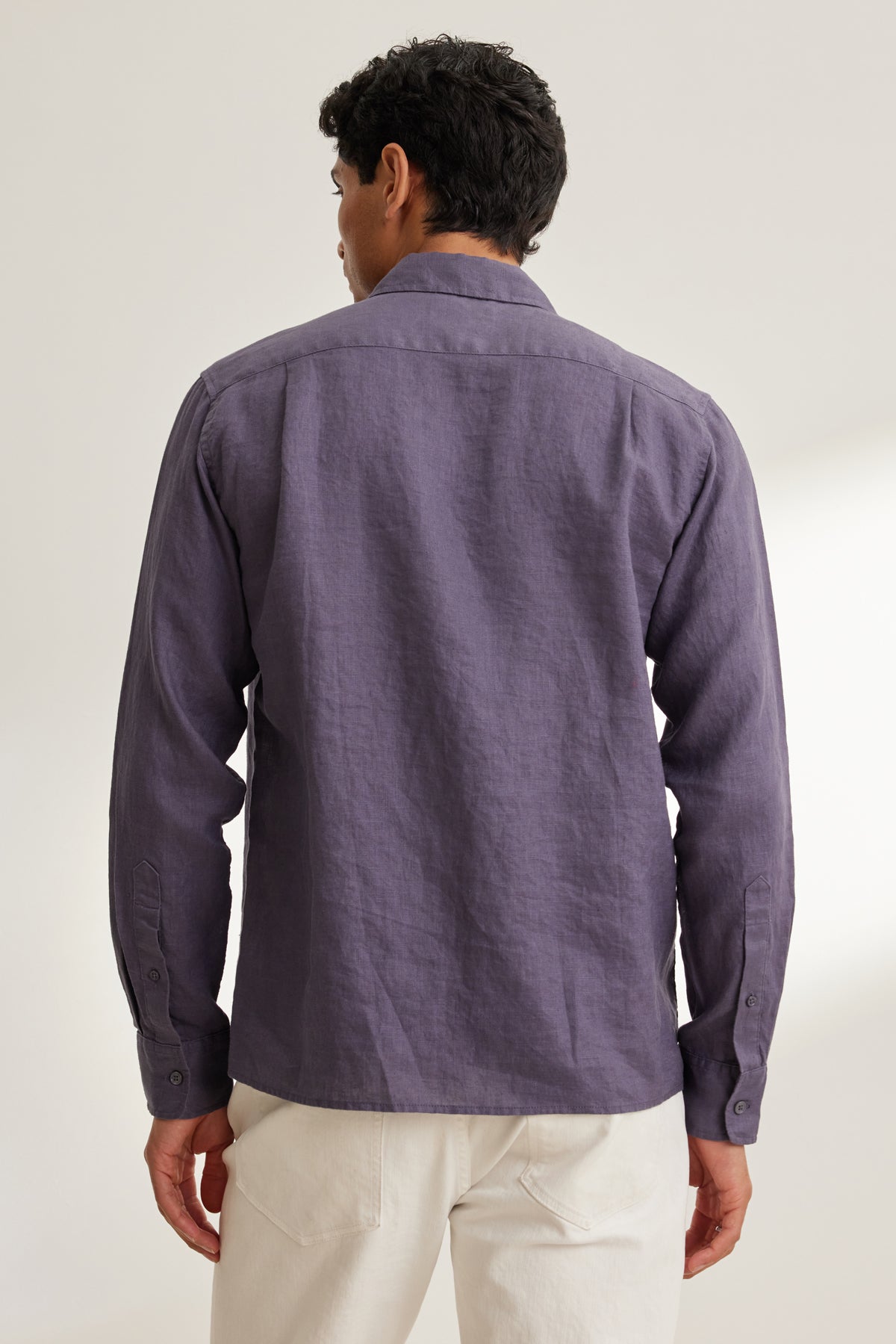   A person stands with their back to the camera, wearing the Velvet by Graham & Spencer CARUSO LINEN BUTTON-UP SHIRT in purple and white pants against a neutral background. 
