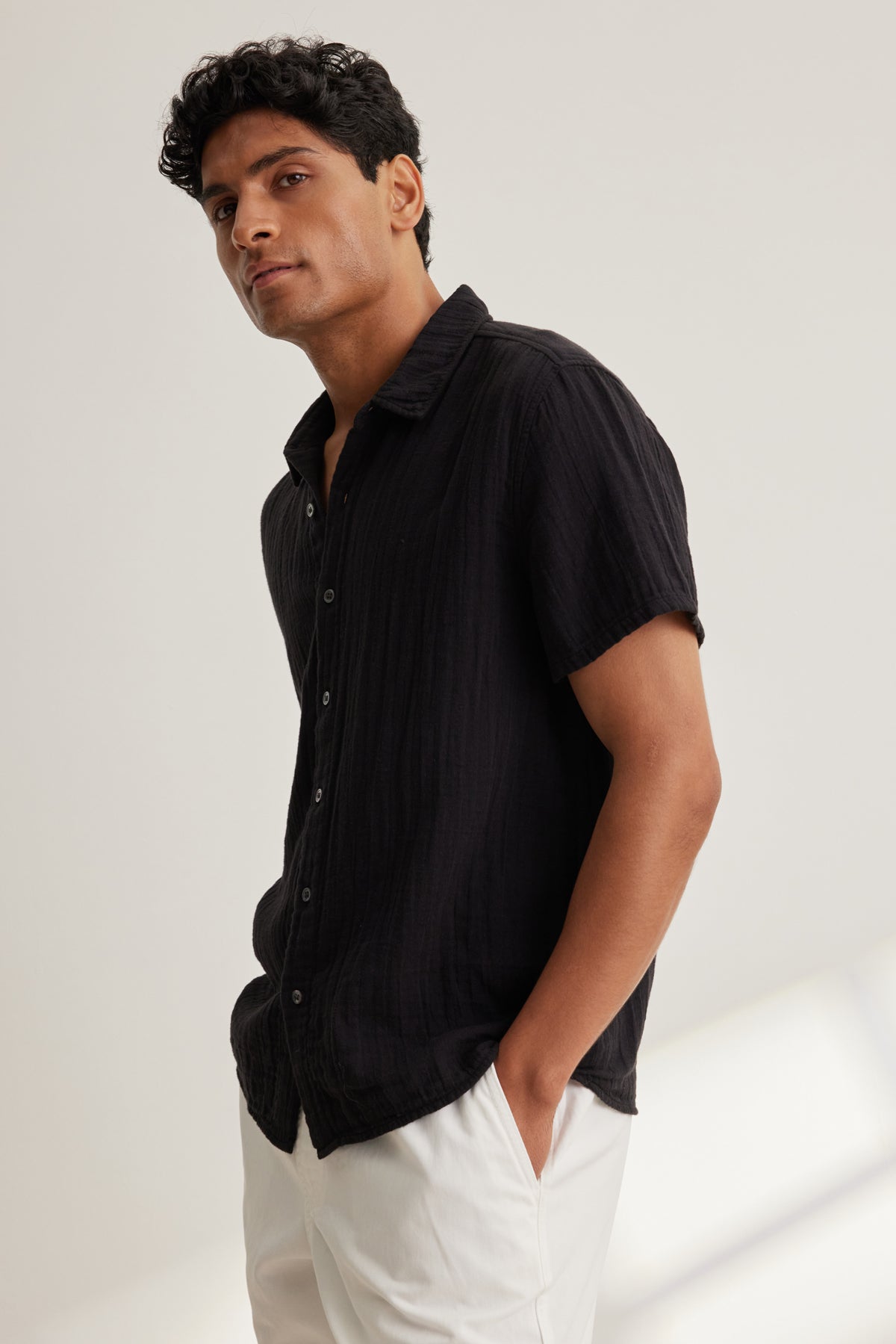   A man stands with his hands in his pockets, wearing the CHRISTIAN COTTON GAUZE BUTTON-UP SHIRT by Velvet by Graham & Spencer—a short-sleeved black cotton gauze shirt—paired with white pants against a plain light background. 