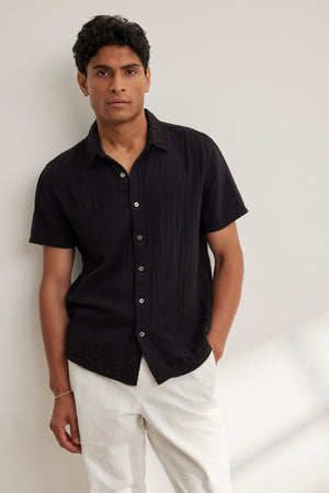 A man stands against a light-colored wall wearing the CHRISTIAN COTTON GAUZE BUTTON-UP SHIRT by Velvet by Graham & Spencer in black, paired with white pants and one hand in his pocket.