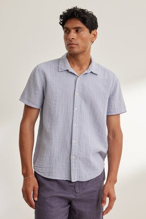 A man wears the Velvet by Graham & Spencer CHRISTIAN COTTON GAUZE BUTTON-UP SHIRT in light blue with dark shorts, standing against a plain, light-colored background.