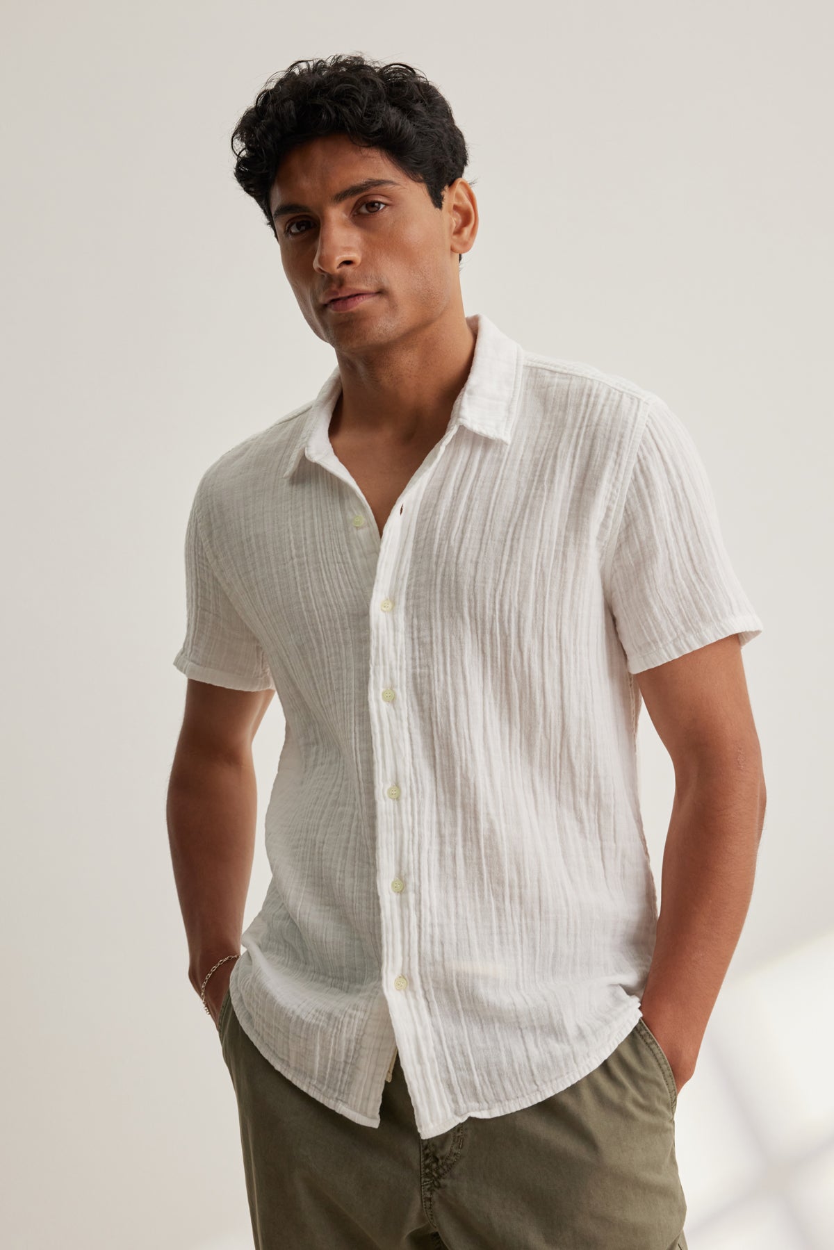   A man stands against a light background, wearing the CHRISTIAN COTTON GAUZE BUTTON-UP SHIRT by Velvet by Graham & Spencer and olive green pants, with his hands in his pockets. 