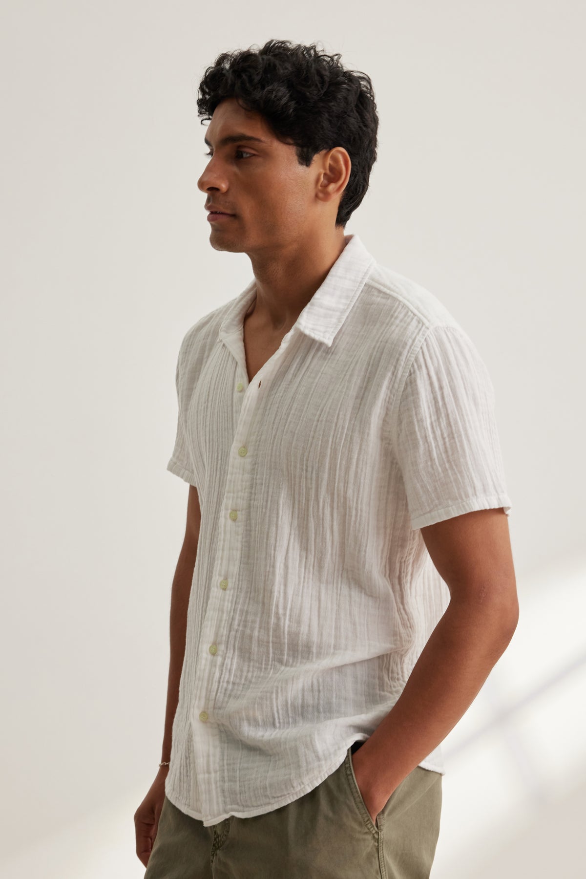 A man stands in profile with his hands in his pockets, wearing the CHRISTIAN COTTON GAUZE BUTTON-UP SHIRT by Velvet by Graham & Spencer and khaki pants against a plain light background.-39152366092481