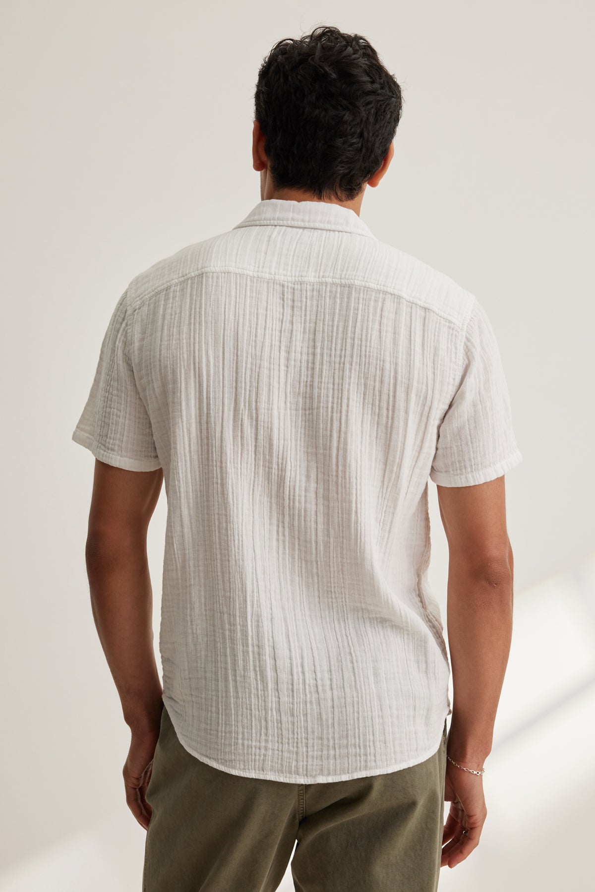   A man faces away from the camera wearing the Velvet by Graham & Spencer CHRISTIAN COTTON GAUZE BUTTON-UP SHIRT in white and olive green pants, set against a plain light background. 