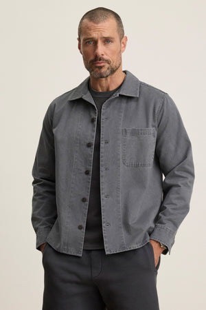 A man with short hair and a trimmed beard wears the CLAY SHIRT JACKET by Velvet by Graham & Spencer—a grey cotton twill jacket with military-style buttons—over a dark tee, hands in pockets, standing against a plain background.
