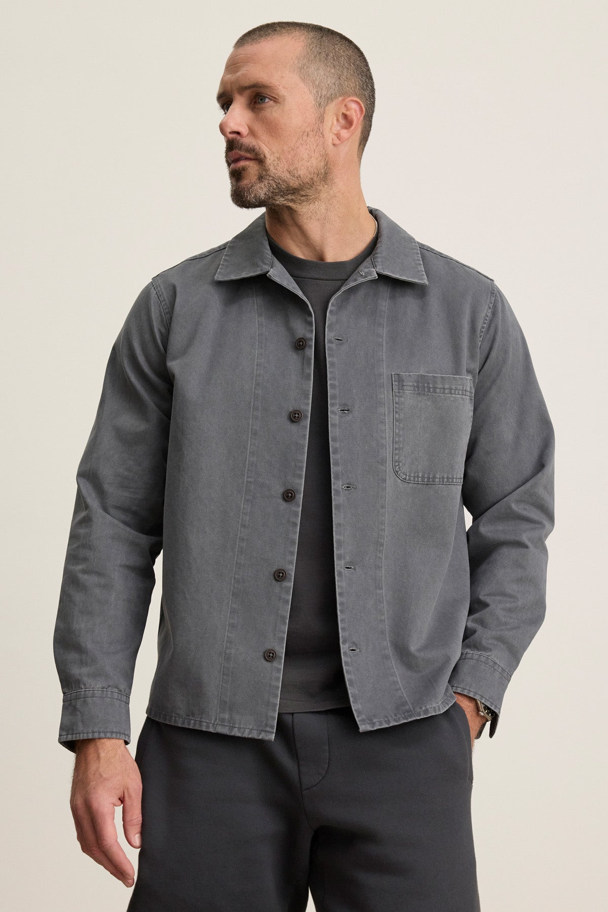 A man in a Velvet by Graham & Spencer CLAY SHIRT JACKET—gray cotton twill with military-style buttons—layered over a dark tee and matching gray pants stands against a plain light background.