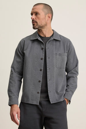 A man in a Velvet by Graham & Spencer CLAY SHIRT JACKET—gray cotton twill with military-style buttons—layered over a dark tee and matching gray pants stands against a plain light background.