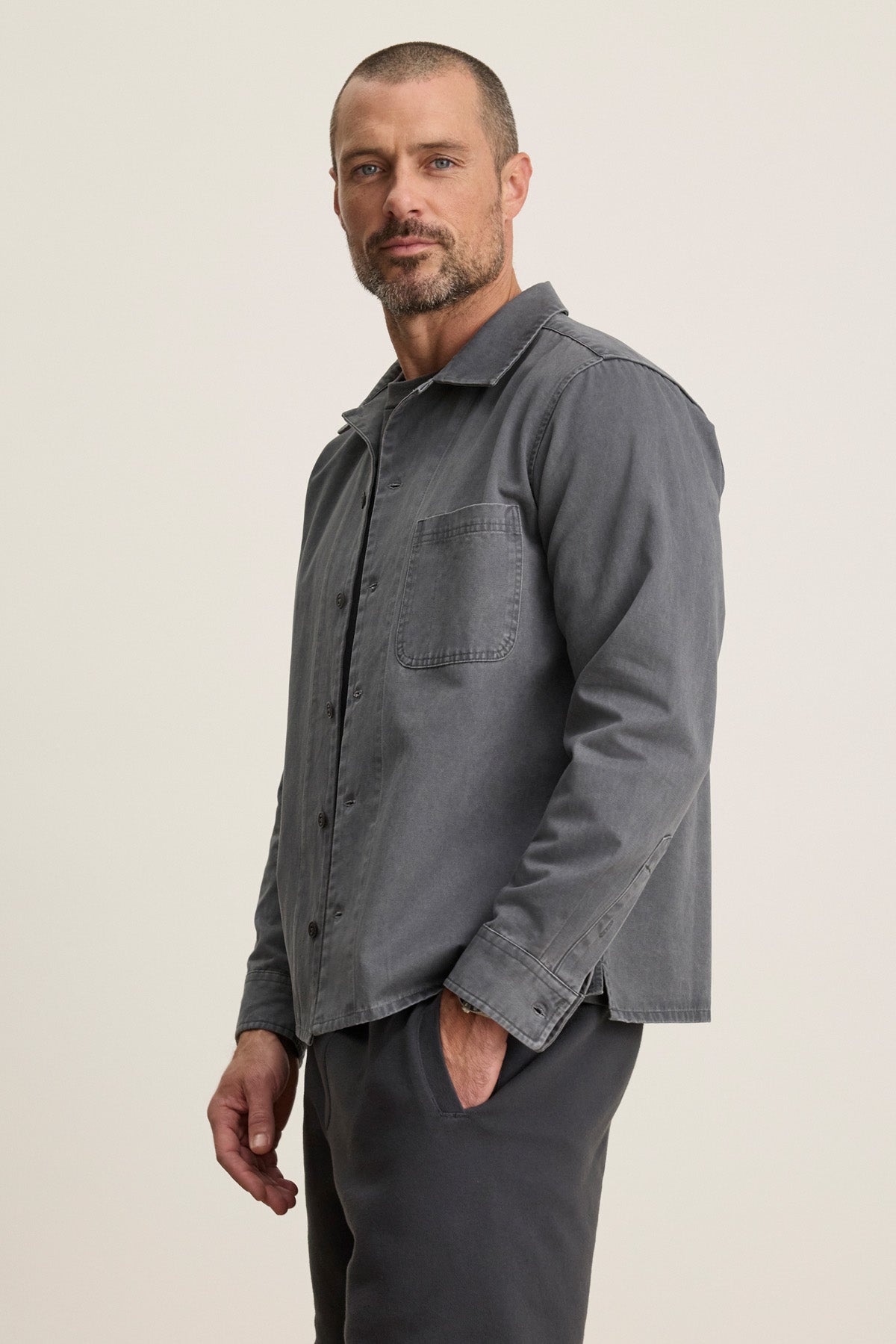 A man with short hair and a trimmed beard models the grey CLAY SHIRT JACKET by Velvet by Graham & Spencer, paired with matching pants, standing with one hand in his pocket against a plain light background.-40452479189185