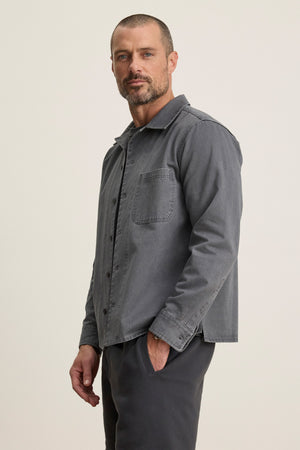 A man with short hair and a trimmed beard models the grey CLAY SHIRT JACKET by Velvet by Graham & Spencer, paired with matching pants, standing with one hand in his pocket against a plain light background.