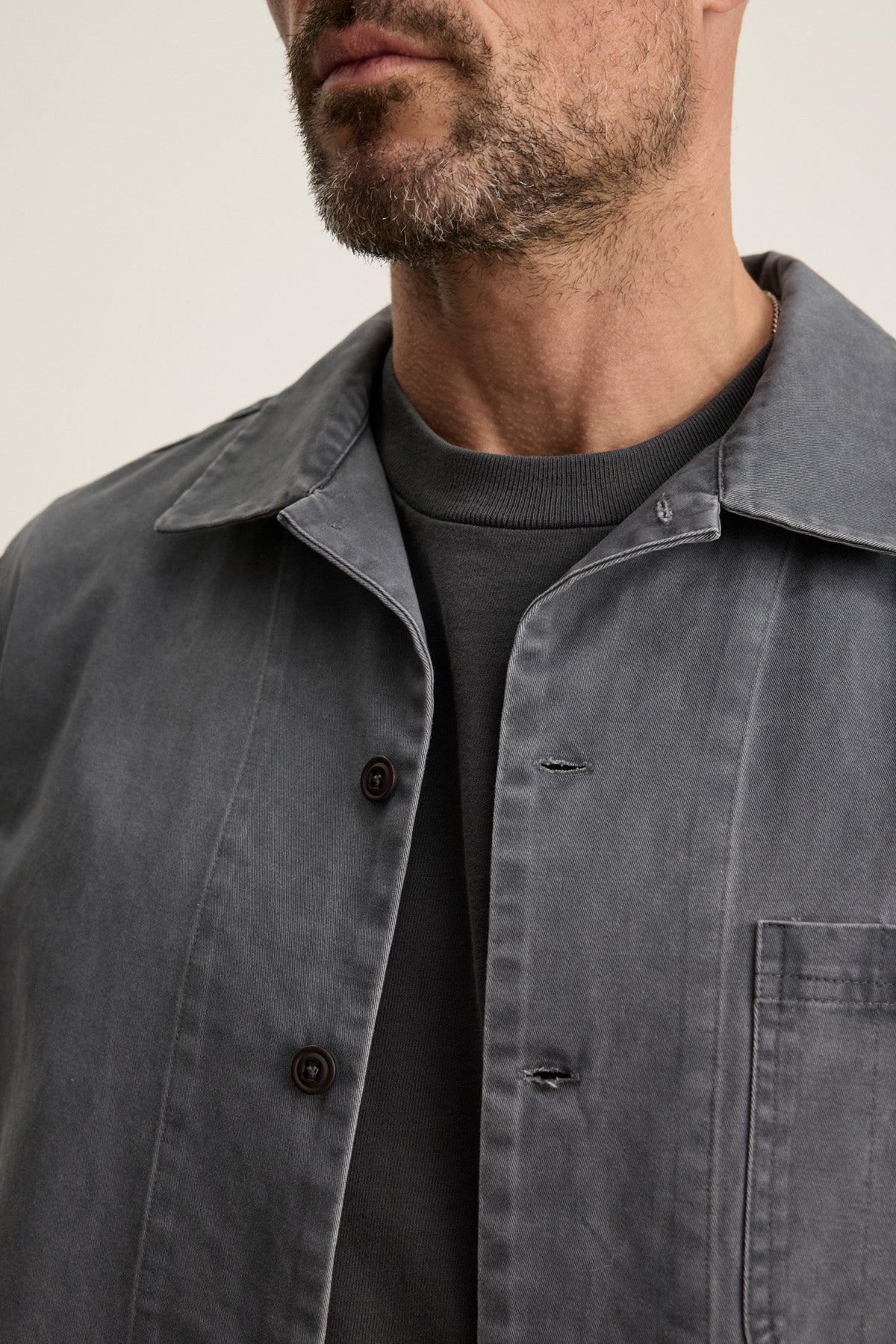 A man with light facial hair wears the CLAY SHIRT JACKET by Velvet by Graham & Spencer, a grey cotton twill piece with military-style buttons, layered over a dark grey crew neck shirt; only his lower face and upper torso are visible.