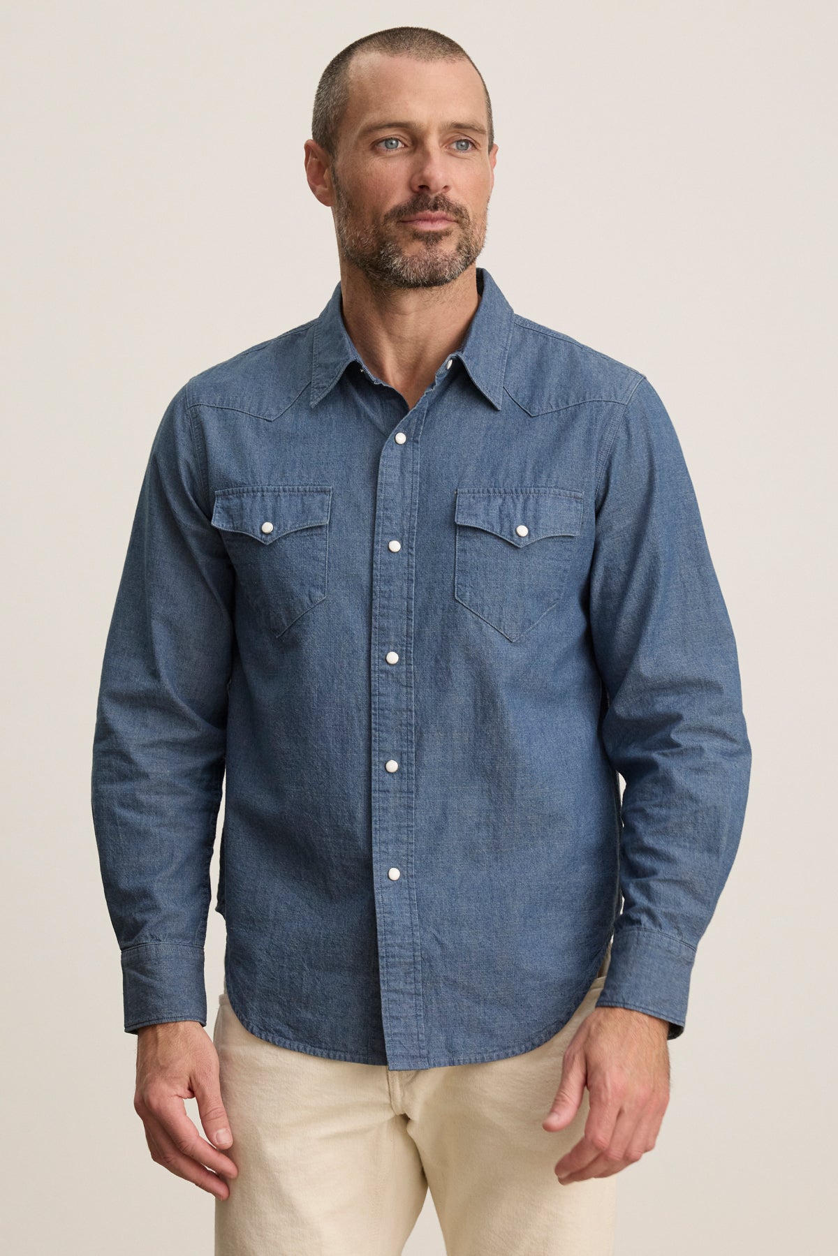 Man wearing the CODY BUTTON-UP SHIRT by Velvet by Graham & Spencer, a tailored fit blue denim shirt with two chest pockets, paired with beige pants, standing against a plain light background.-39934272602305