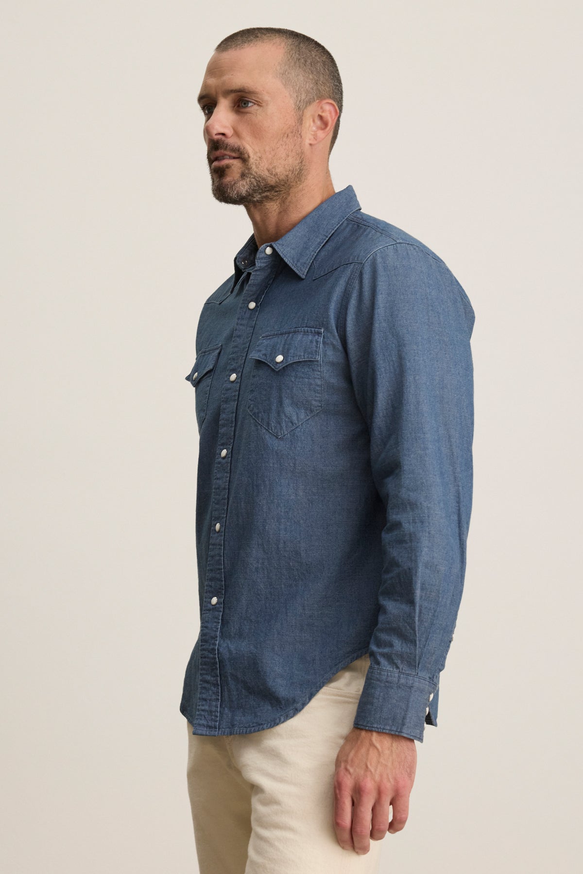 A man with short hair and a beard wears the Velvet by Graham & Spencer CODY BUTTON-UP SHIRT, a tailored Western-style blue denim shirt, paired with light pants, standing against a plain background.-39934272635073