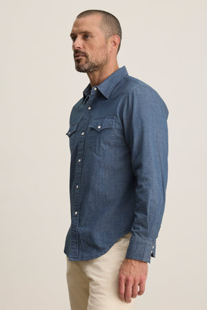 A man with short hair and a beard wears the Velvet by Graham & Spencer CODY BUTTON-UP SHIRT, a tailored Western-style blue denim shirt, paired with light pants, standing against a plain background.