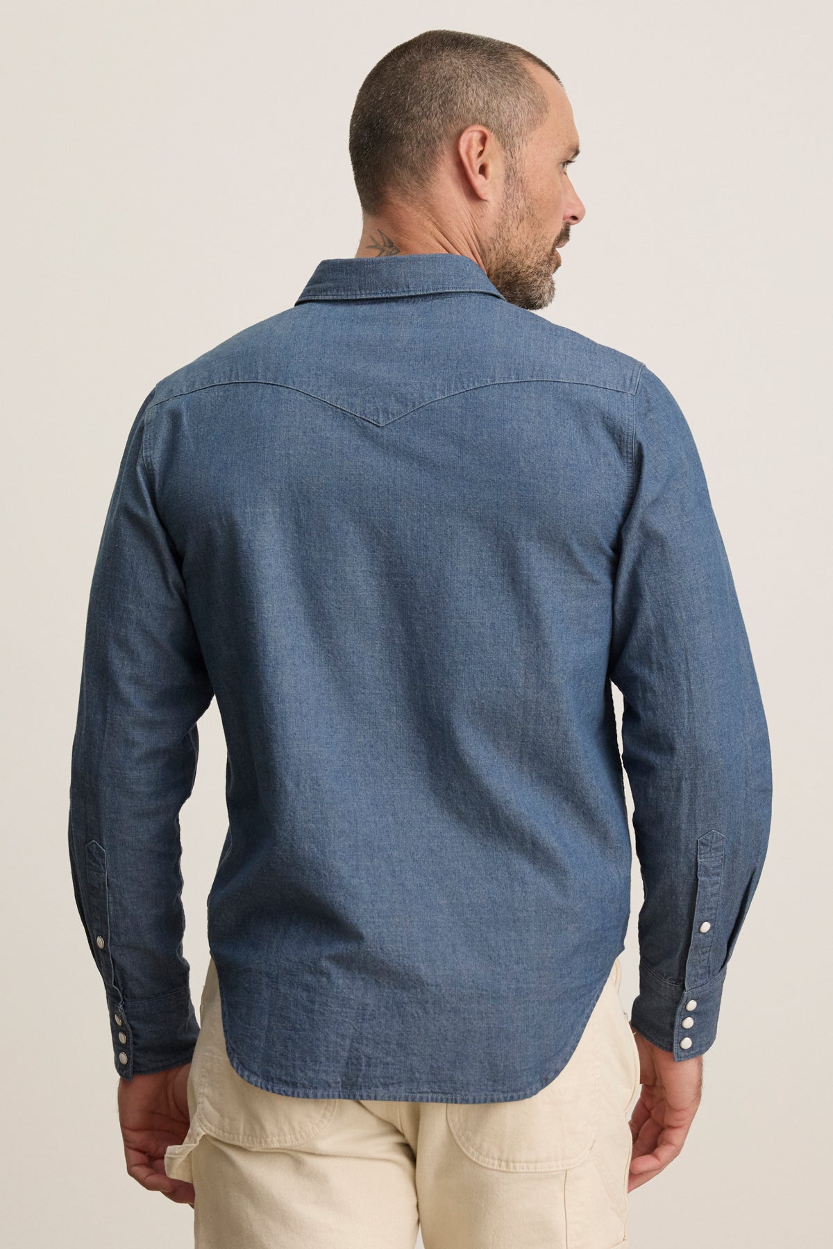 A man with short hair is seen from the back, wearing the Velvet by Graham & Spencer CODY BUTTON-UP SHIRT in blue denim and light-colored pants against a plain background.-39934272700609