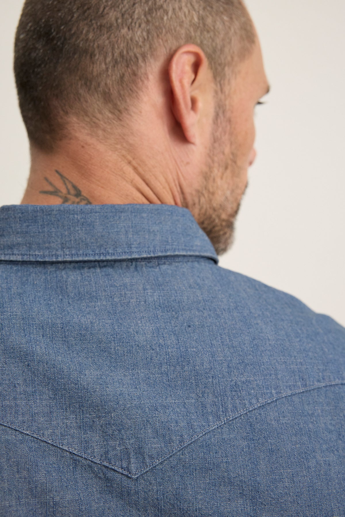 A man seen from behind wears the Velvet by Graham & Spencer CODY BUTTON-UP SHIRT in blue denim with a tailored fit; a tattoo is partially visible on the back of his neck.-39934272897217