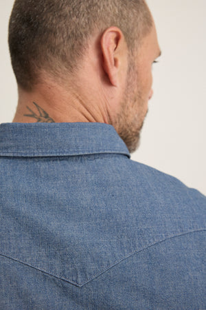 A man seen from behind wears the Velvet by Graham & Spencer CODY BUTTON-UP SHIRT in blue denim with a tailored fit; a tattoo is partially visible on the back of his neck.