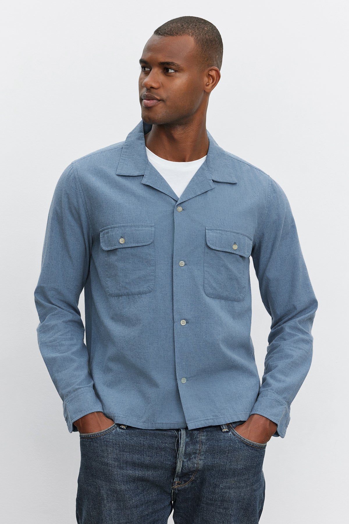 A man is standing in the DONOVAN BUTTON-UP SHIRT by Velvet by Graham & Spencer with his hands in his pockets. He is looking off to the left against a plain white background, showcasing the refined style of his camp collar cotton blend twill garment.-37893886935233