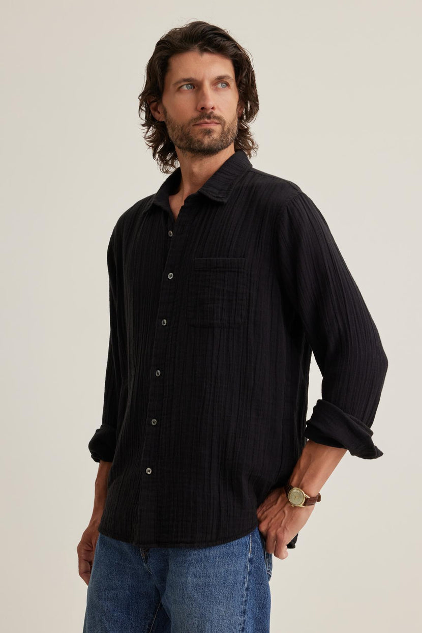 A man with long hair wearing the Velvet by Graham & Spencer ELTON Cotton Gauze Button-Up Shirt and blue jeans looks to the side.