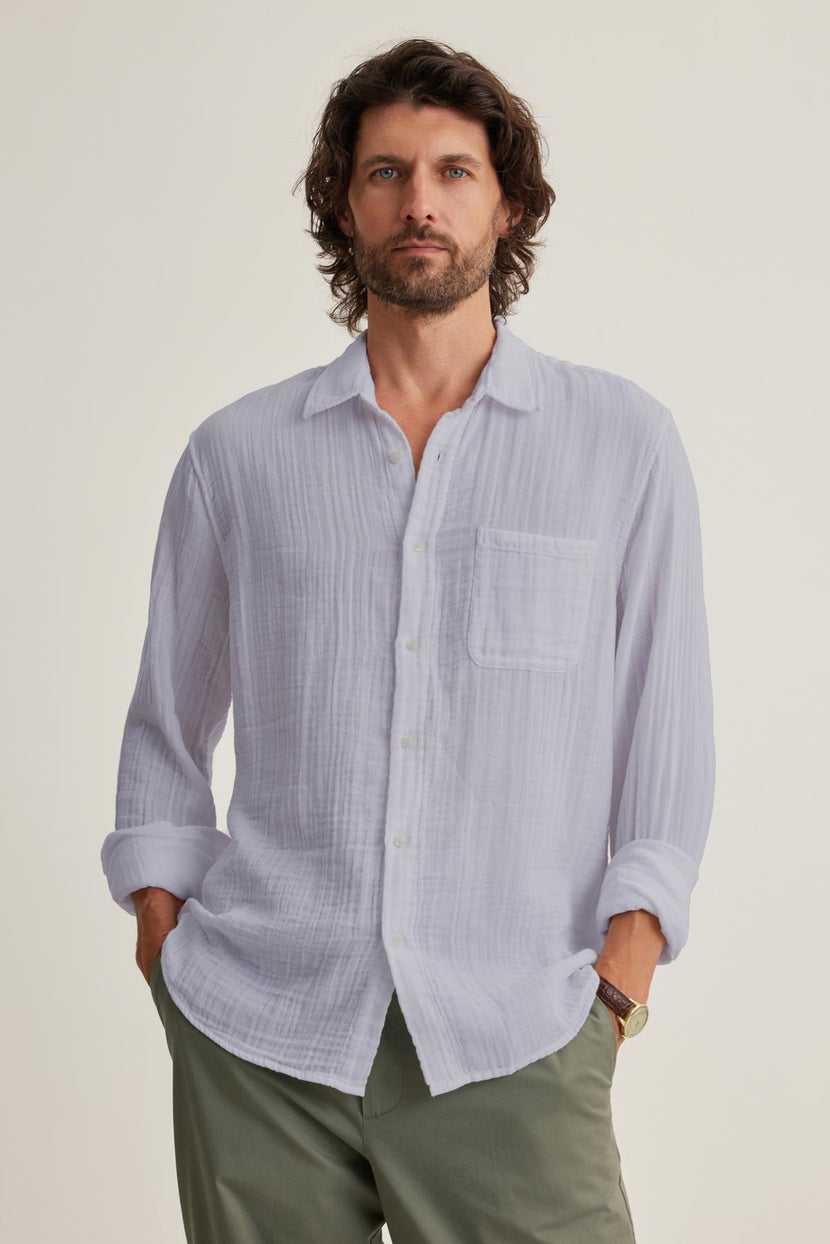 A man with curly hair stands with hands in pockets, wearing the ELTON COTTON GAUZE BUTTON-UP SHIRT by Velvet by Graham & Spencer and green pants—a versatile choice.