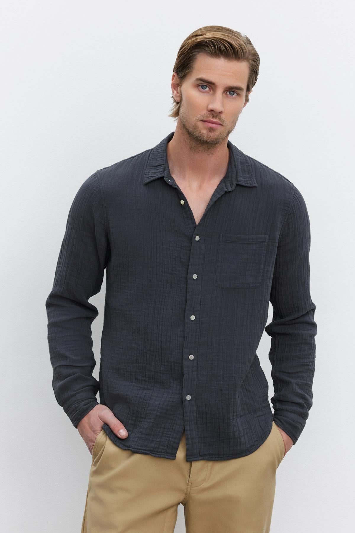   A person stands against a plain background, wearing the ELTON COTTON GAUZE BUTTON-UP SHIRT by Velvet by Graham & Spencer and tan pants. With hands in pockets and a neutral expression, this outfit presents a versatile choice for various occasions. 