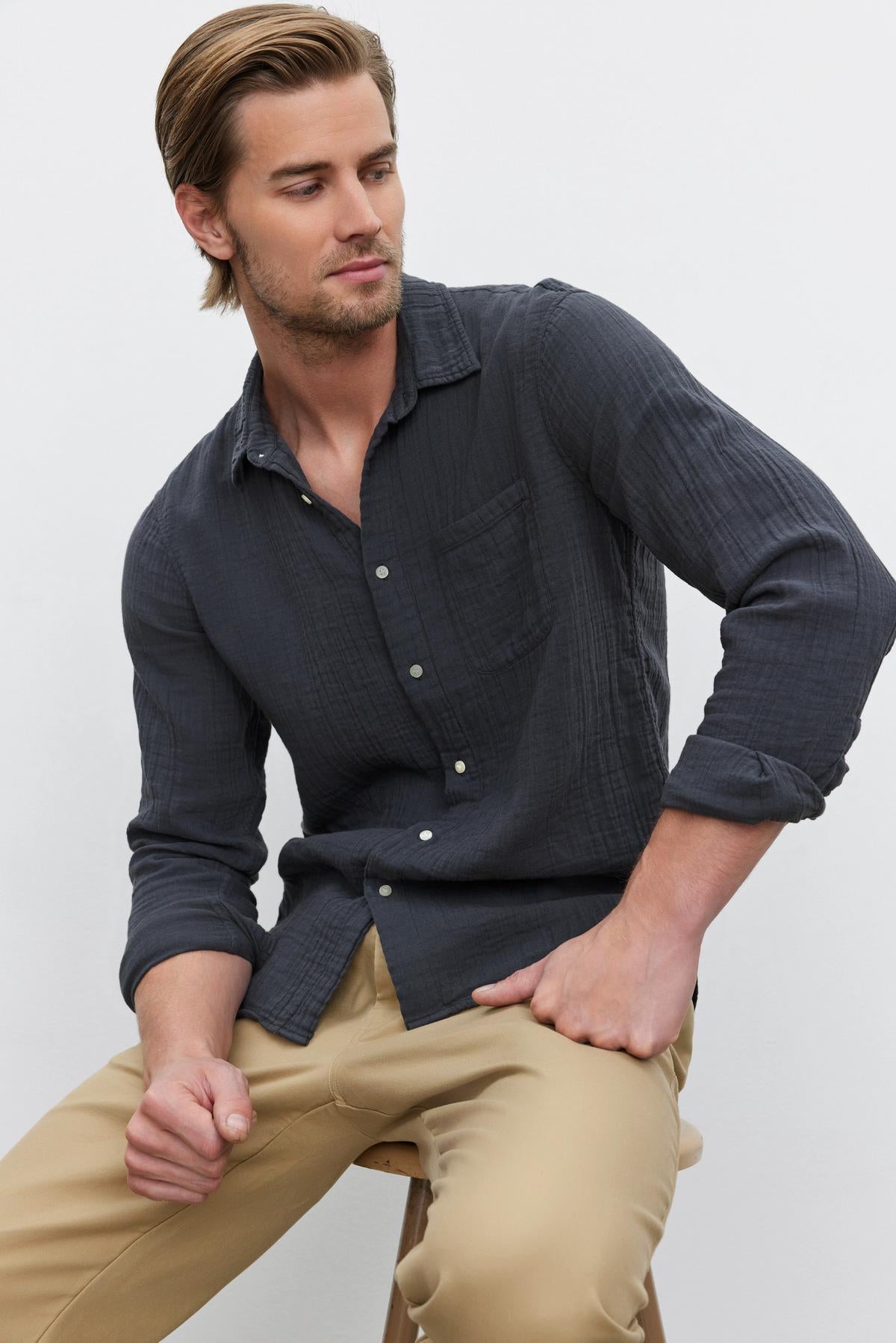   A person with neatly styled hair wearing a dark ELTON COTTON GAUZE BUTTON-UP SHIRT by Velvet by Graham & Spencer and beige pants sits on a stool against a plain white background—a versatile choice for any occasion. 