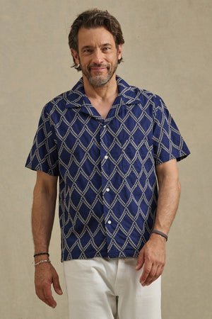 A man with brown hair and a beard wears the Velvet by Graham & Spencer ESTEBAN SHIRT—a navy camp collar shirt with a white geometric pattern—and white pants, standing against a plain background.