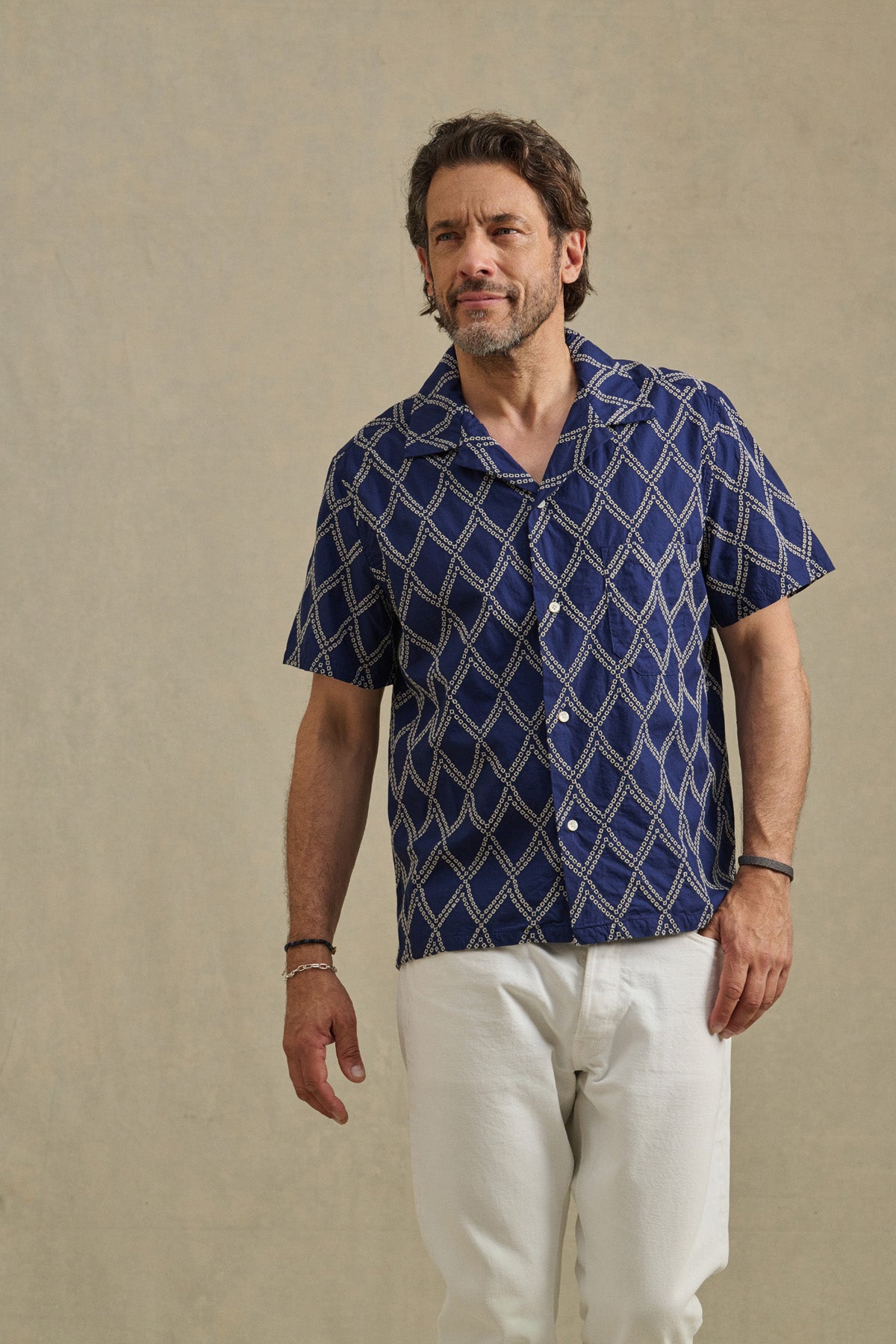 Man with short hair and beard wears the ESTEBAN SHIRT by Velvet by Graham & Spencer—a blue patterned camp collar style—paired with white pants, standing against a plain beige background.-41626552631489