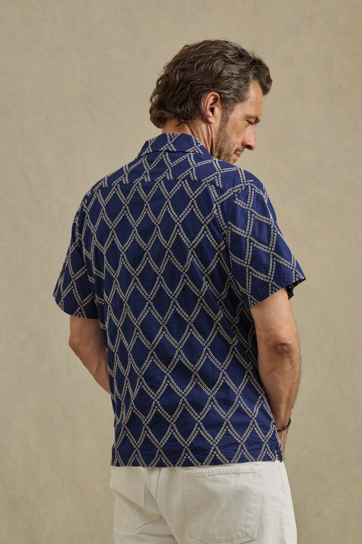 A man faces away wearing the ESTEBAN SHIRT by Velvet by Graham & Spencer—a short-sleeve navy camp collar shirt with a white geometric pattern—and white pants, standing against a plain beige background.-41626552762561