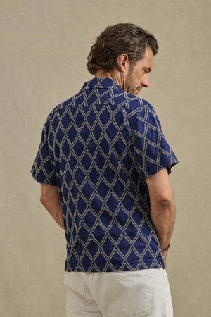 A man faces away wearing the ESTEBAN SHIRT by Velvet by Graham & Spencer—a short-sleeve navy camp collar shirt with a white geometric pattern—and white pants, standing against a plain beige background.