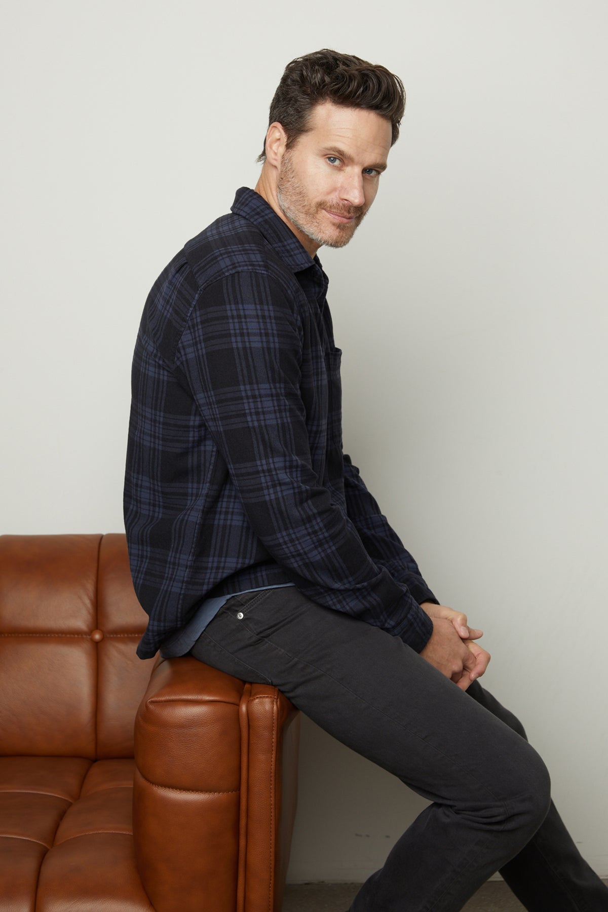   A man in a warm Velvet by Graham & Spencer FREDDY PLAID SHIRT sitting on a brown leather couch. 