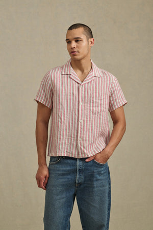 A person wears the HOBBS SHIRT by Velvet by Graham & Spencer—a short-sleeve, pink and white striped button-up—paired with blue jeans, standing against a plain beige background.