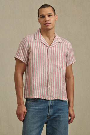 A person wears the HOBBS SHIRT by Velvet by Graham & Spencer— a short-sleeve, button-up with pink and white vertical stripes—paired with blue jeans, standing against a neutral background.
