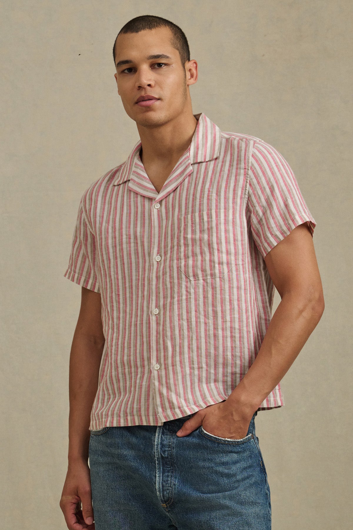 Someone wearing the HOBBS SHIRT by Velvet by Graham & Spencer—a pink and white striped, short-sleeve button-up—is standing against a plain beige background, paired with blue jeans.-41374390517953