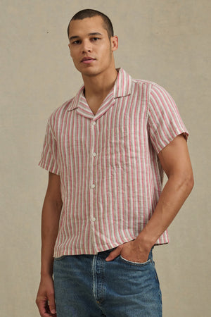 Someone wearing the HOBBS SHIRT by Velvet by Graham & Spencer—a pink and white striped, short-sleeve button-up—is standing against a plain beige background, paired with blue jeans.