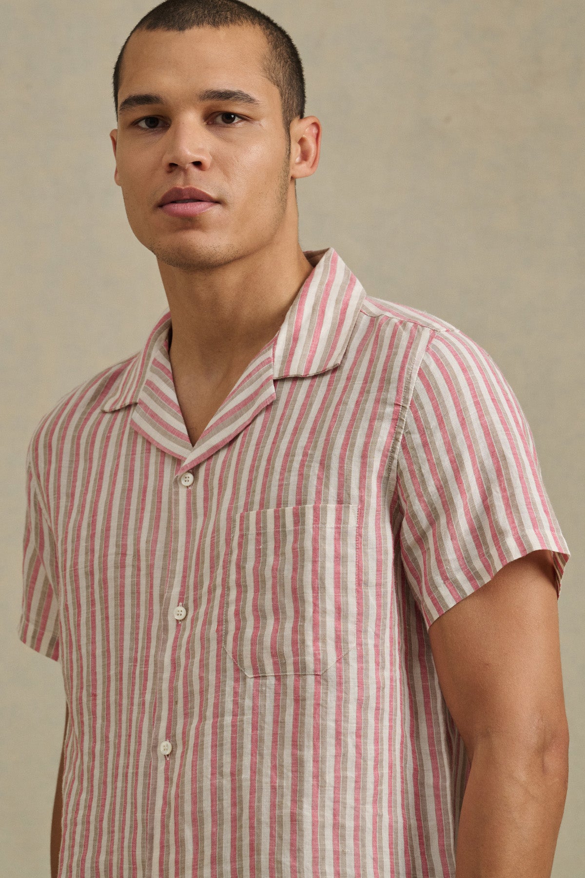 A man wears the HOBBS SHIRT by Velvet by Graham & Spencer, a short-sleeve, button-up featuring pink, white, and beige vertical stripes, standing against a neutral background.