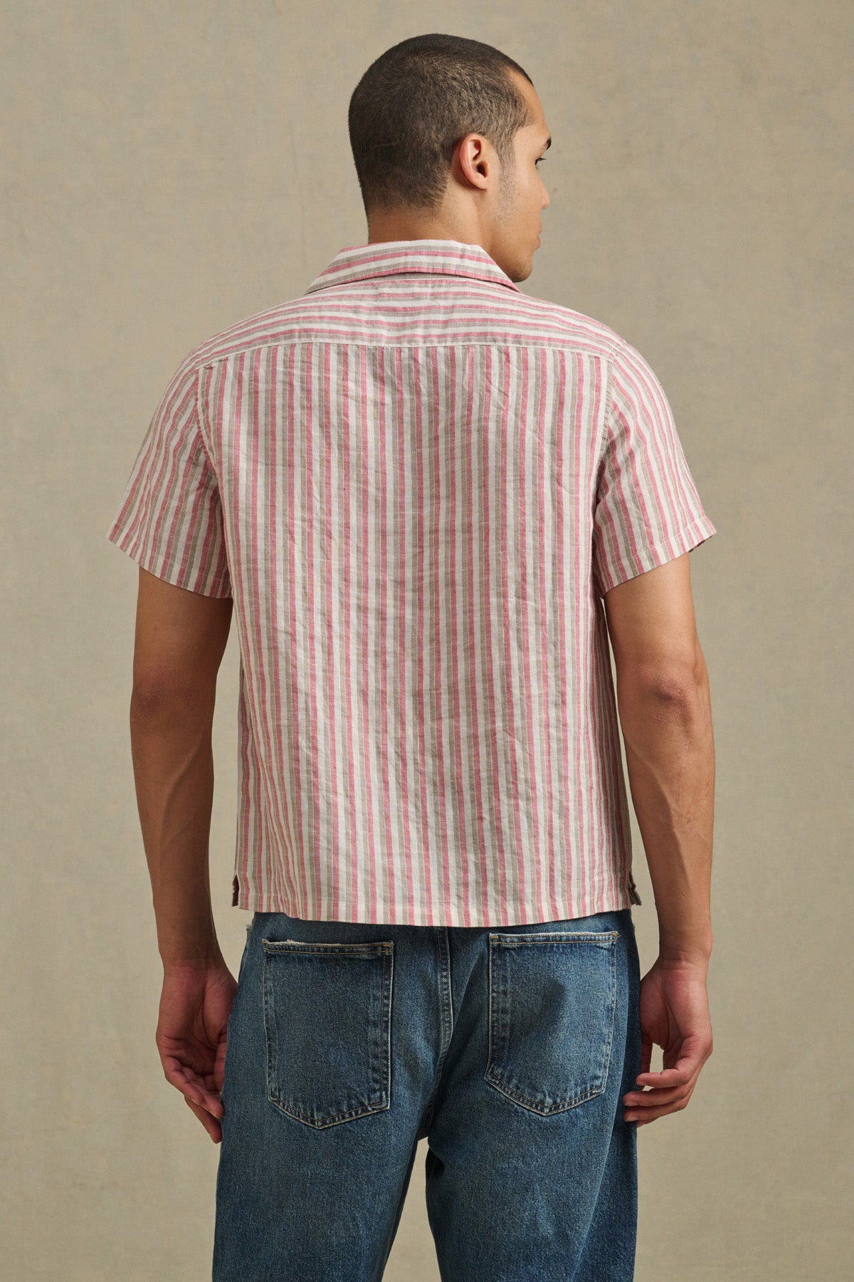 A man stands facing away, wearing the HOBBS SHIRT by Velvet by Graham & Spencer—a short-sleeve, red and white vertical striped shirt—paired with blue jeans against a neutral background.