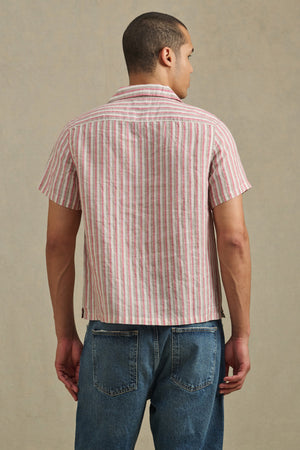A man stands facing away, wearing the HOBBS SHIRT by Velvet by Graham & Spencer—a short-sleeve, red and white vertical striped shirt—paired with blue jeans against a neutral background.