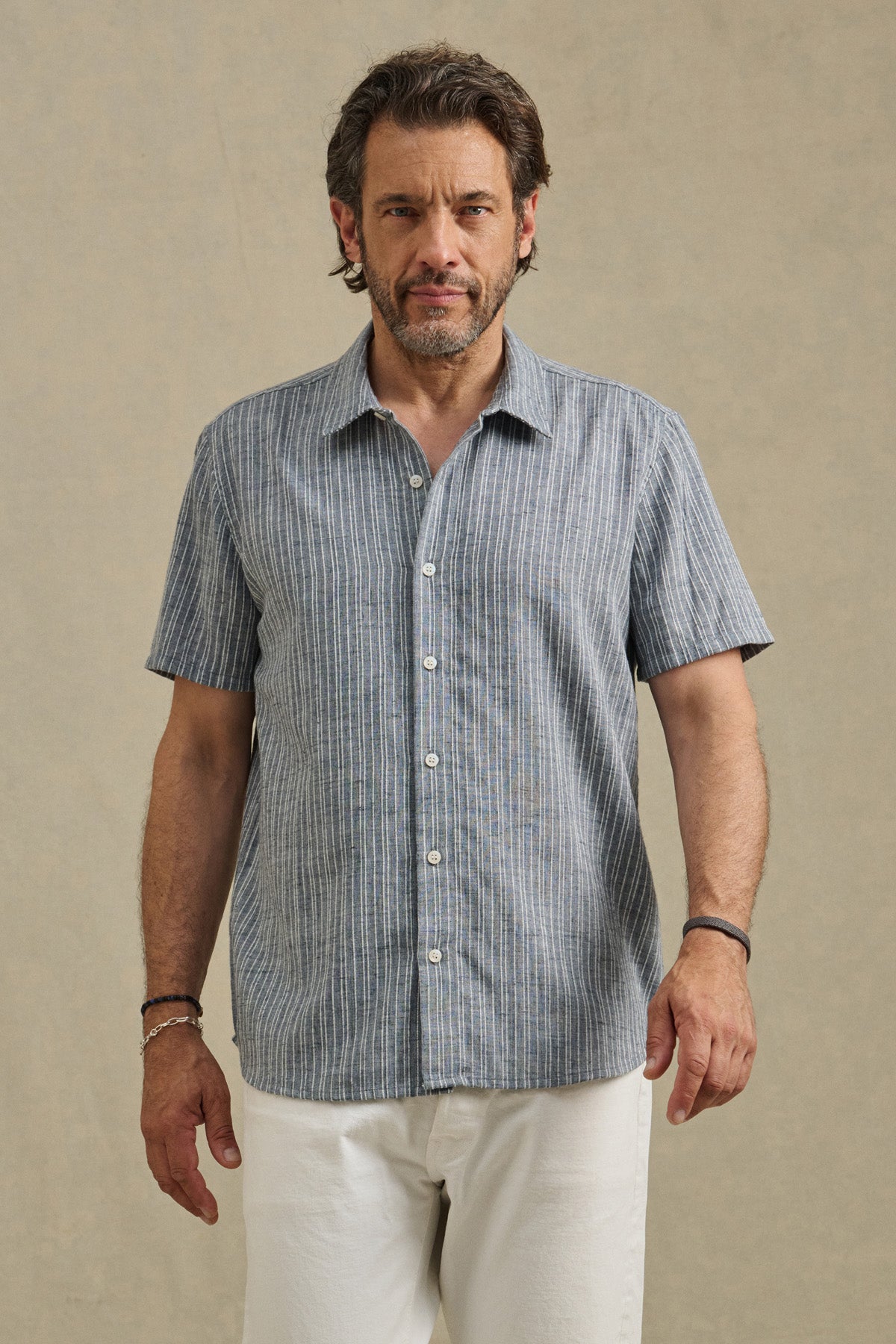 A man faces forward in the KAIDEN SHIRT by Velvet by Graham & Spencer, a short-sleeve blue and white striped cotton shirt with a classic collar, paired with white pants against a plain beige background.-41548219384001