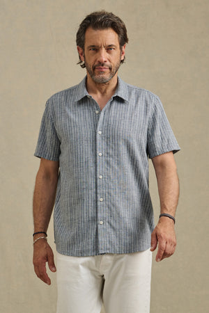 A man faces forward in the KAIDEN SHIRT by Velvet by Graham & Spencer, a short-sleeve blue and white striped cotton shirt with a classic collar, paired with white pants against a plain beige background.