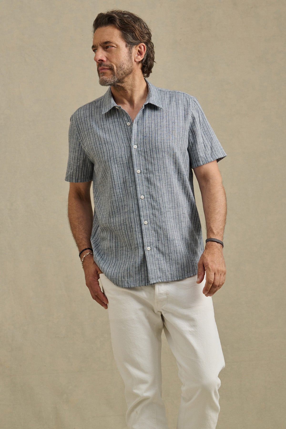 A man wears the KAIDEN SHIRT by Velvet by Graham & Spencer, a classic-collar, short-sleeve striped cotton button-up, paired with white pants against a plain beige background.-41548219416769