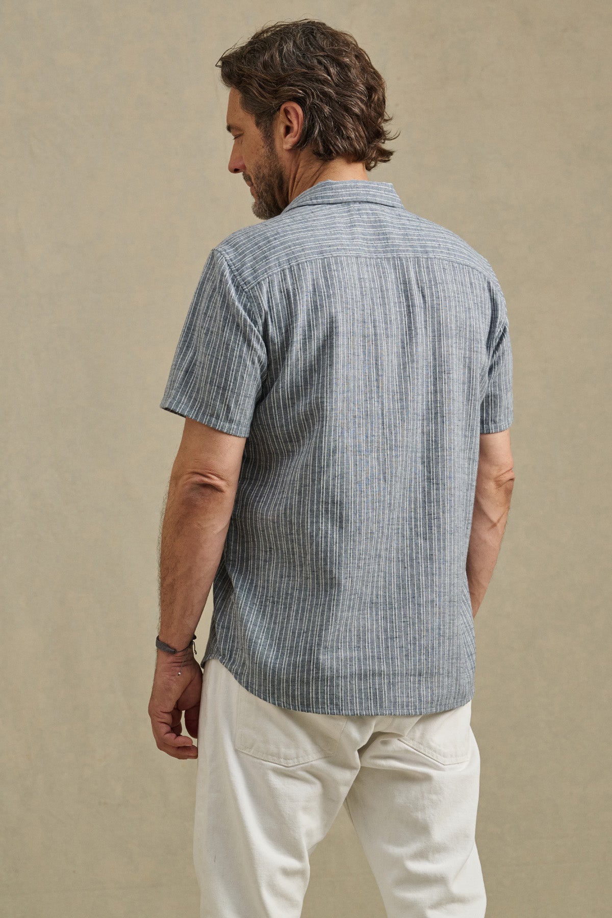 A man faces away, wearing the KAIDEN SHIRT by Velvet by Graham & Spencer—a blue striped cotton button-up with a classic collar—paired with white pants against a plain beige background.-41548219547841