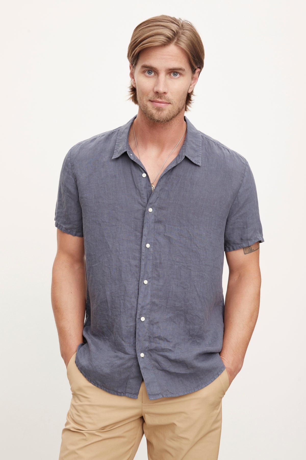  A man with shoulder-length hair stands with his hands in his pockets, wearing a short-sleeve MACKIE LINEN BUTTON-UP SHIRT by Velvet by Graham & Spencer and tan pants, perfect for a summer wardrobe. 