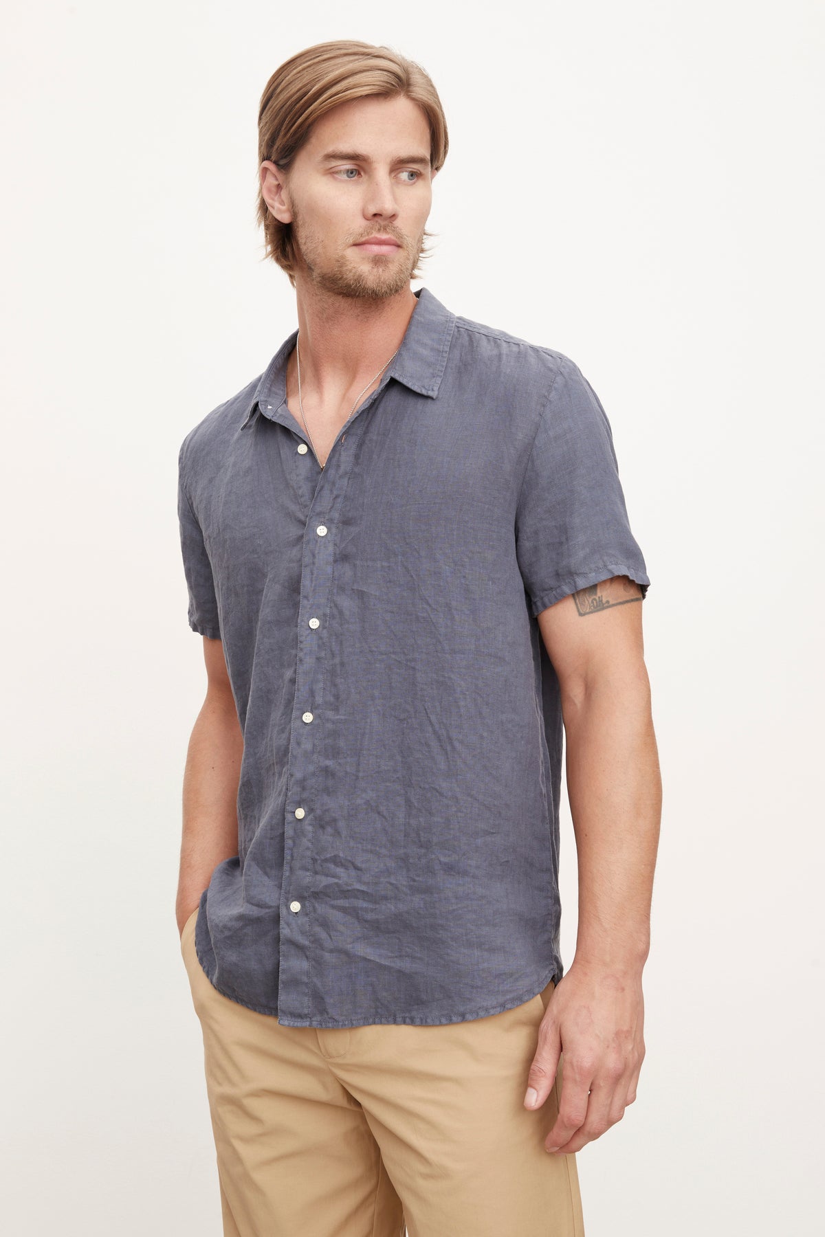 A person with light brown hair is wearing a short-sleeved blue MACKIE LINEN BUTTON-UP SHIRT by Velvet by Graham & Spencer and beige pants, standing against a plain white background, showcasing their perfect summer wardrobe.-36918688022721