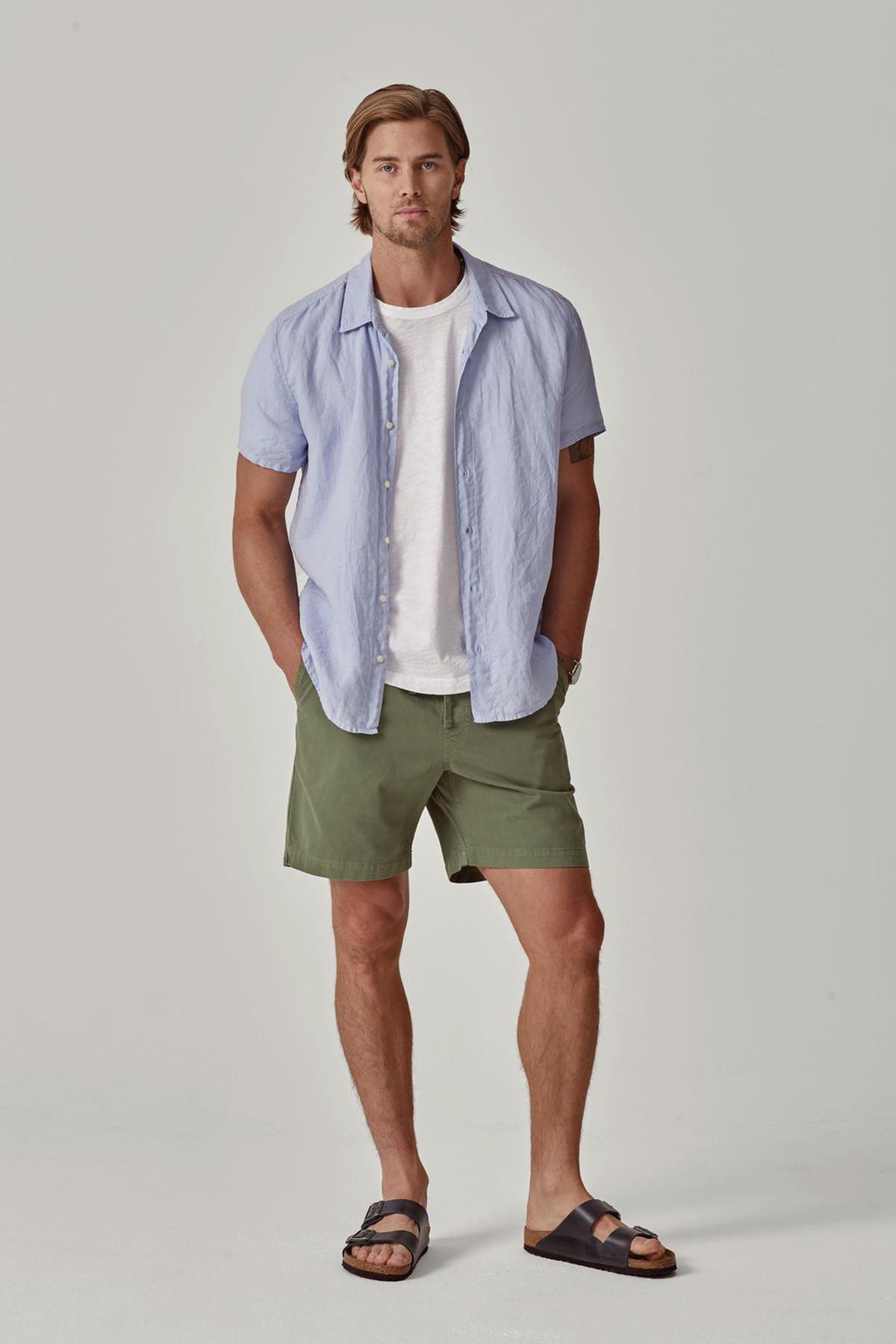 A man with shoulder-length hair stands wearing a Velvet by Graham & Spencer MACKIE LINEN BUTTON-UP SHIRT over a white t-shirt, green shorts, and black sandals, hands in pockets.-36753546281153